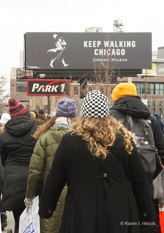 Marchers walking down Chicago street. Above is a Johnny Walker billboard saying Keep Walking Chicago